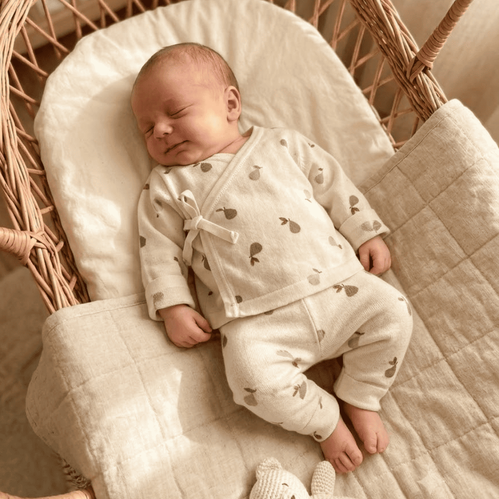 Nouveau-né dormant paisiblement dans un berceau en rotin, vêtu d'un ensemble naissance deux pièces en velours de coton écru imprimé petites poires avec fermeture cache-cœur.