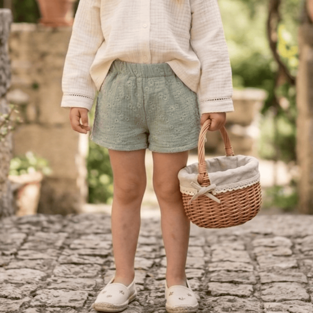 Petite fille portant un short vert sauge brodé en gaze de coton avec panier en osier, tenue estivale enfant.